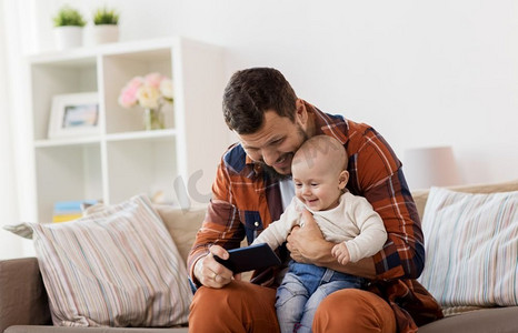 家庭、父母和人的概念-快乐的父亲和家里有智能手机的小男孩。父亲和男婴在家里带着智能手机