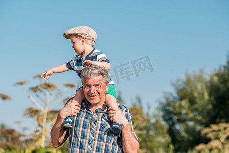 祖父将孙子蹒跚学步的男孩扛在肩上。一个骑在爷爷S背上的孩子回到了户外。