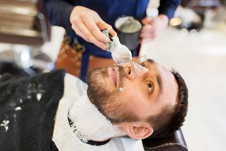 美容和人的概念-男人和理发师用刷子在理发店将剃须泡沫应用到胡须上。男人和理发师在胡须上涂剃须泡沫