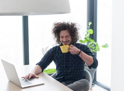 一个年轻人一边喝着咖啡，一边在家里用笔记本电脑工作