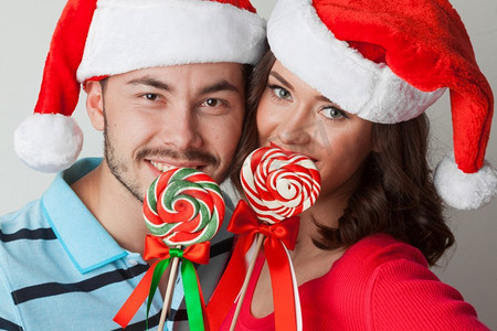 拿着棒棒糖的情侣摄影照片_拿着棒棒糖的圣诞情侣年轻的快乐有趣的夫妇在圣诞节圣诞老人帽子与棒棒糖