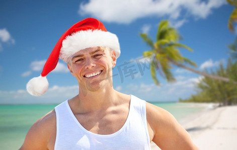 年轻人海边摄影照片_圣诞节、节假日和人们的概念快乐--海滩上戴着圣诞帽的年轻人微笑着。圣诞快乐的年轻人戴着圣诞帽在海滩上