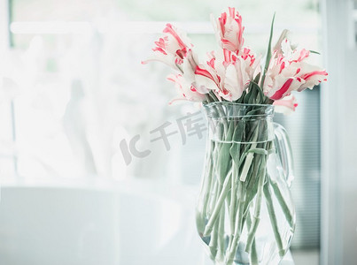 白色窗边摄影照片_窗边白色桌子上的玻璃罐里放着一束漂亮的郁金香。室内设计中的花卉。舒适的家。春天到了