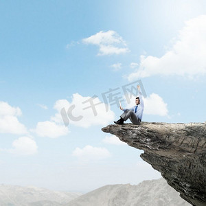 云层上摄影照片_站在顶端。年轻的商人坐在岩石的边缘