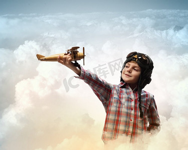 戴着飞行员帽的小男孩S。戴着飞行员头盔的小男孩在云的背景下玩玩具飞机