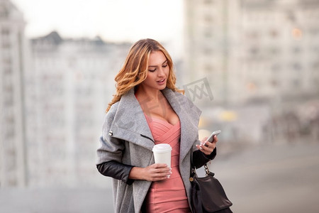城市景象摄影照片_一名女商人通过手机查看电子邮件，手持咖啡杯，与城市景象相映衬。