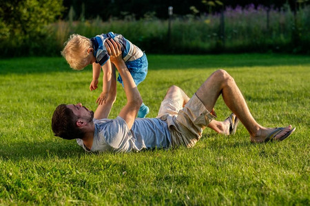 草地玩的孩子摄影照片_快乐的男人和孩子在草地上享受户外乐趣。公园里父子俩在绿茵茵的草地上休息的家庭生活场景。