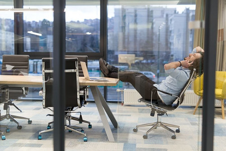 这是放松的时候。疲惫的年轻休闲商人在办公室的办公桌前放松