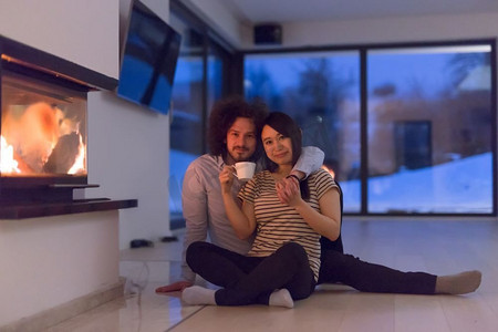 惊恐聊天表情摄影照片_年轻浪漫的多民族情侣坐在家里壁炉前的地板上，在寒冷的冬夜里聊天喝茶