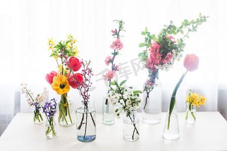 瓶子里的花束摄影照片_靠窗的桌子上放着水，瓶子里放着不同的美丽的花。桌上摆着一群不同的花