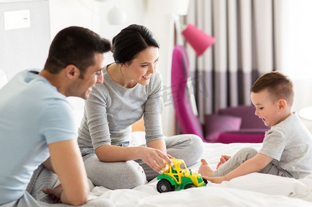 小汽车玩具摄影照片_人和家庭的概念-快乐的孩子带着玩具拖拉机，父母在家里或酒店房间的床上玩耍。幸福的家庭在家里或酒店房间的床上