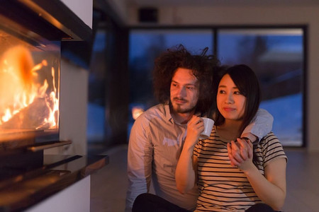 年轻浪漫的多民族情侣坐在家里壁炉前的地板上，在寒冷的冬夜里聊天喝茶
