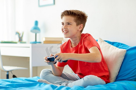 孩子电子游戏摄影照片_游戏，技术和人的概念-微笑的男孩，带着游戏手柄在家玩电子游戏。带着游戏手柄在家玩电子游戏的快乐男孩