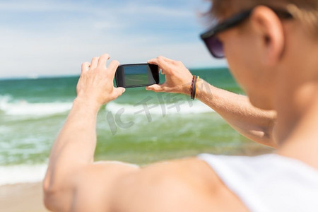 夏日拍照摄影照片_暑假和人的概念-年轻人在海滩上用智能手机拍摄大海。一名男子在夏日海滩用智能手机拍照
