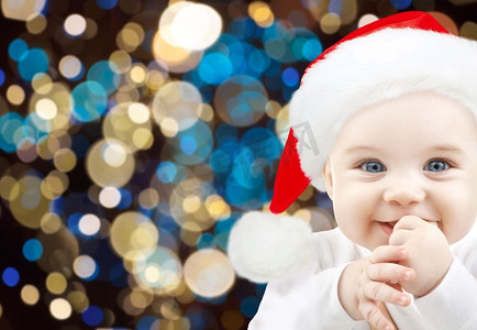 圣诞宝宝摄影照片_节日，童年和人们的概念-快乐的婴儿在圣诞老人的帽子上灯光背景。圣诞彩灯下戴着圣诞帽的快乐宝宝