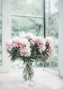 有窗背景摄影照片_窗边地板上的玻璃花瓶里有一束可爱的粉色牡丹。室内设计中的花卉。舒适的家。