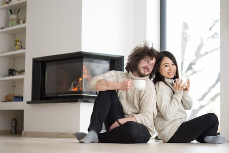 年轻浪漫的多民族情侣坐在家里壁炉前的地板上，在寒冷的冬日聊天喝咖啡