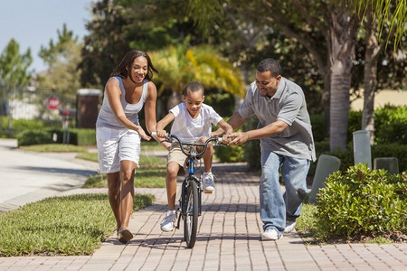 男孩骑摄影照片_一个年轻的非裔美国家庭，有一个男孩骑着自行车，他高兴而兴奋的父母在他旁边鼓励他