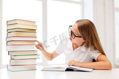 学生一堆书摄影照片_教育、学校和人的概念-戴眼镜的学生女孩，桌上坐着一堆书。戴眼镜的女学生在学校看书