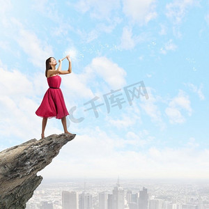吹裙子摄影照片_吹横笛的女人。年轻女子在红色礼服上的岩石边缘演奏横笛