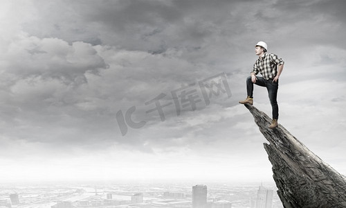 重回巅峰摄影照片_年轻自信的建设者站在高高的城市上方的岩石边。攻克巅峰