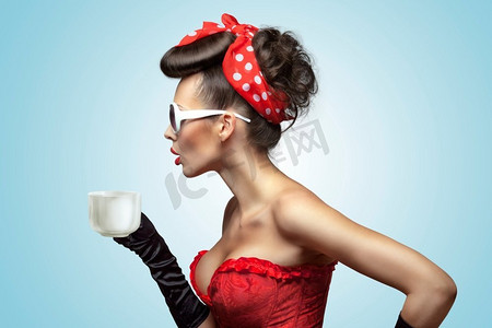这张复古照片中，迷人的美女戴着复古手套，头发上别着红丝带，手里拿着一杯热咖啡或茶，然后冷却。