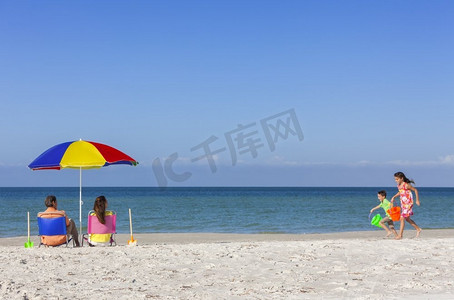 打伞男人背影摄影照片_在阳光明媚的海滩上，一个幸福的家庭，父母、女儿和儿子在帆布椅上打伞玩耍的背影