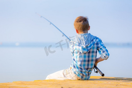 穿蓝衬衫的男孩坐在馅饼上。穿着蓝色衬衫的男孩坐在海边的码头上，手里拿着钓鱼竿