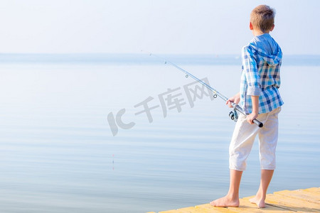 穿着蓝色衬衫的男孩站在海边的码头上，手里拿着一根钓鱼竿