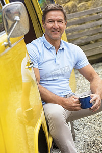 英俊、成功、快乐的中年男子坐在一辆黄色露营面包车的门口，用锡杯或杯子喝茶或咖啡