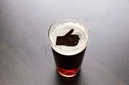 喜欢新鲜啤酒。在黑色桌子上的啤酒杯泡沫上竖起大拇指的剪影，从上面看。