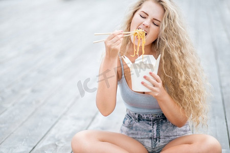 幸福的年轻女子留着长长的卷发，拿着午餐盒，用筷子吃着中式外卖的面条。