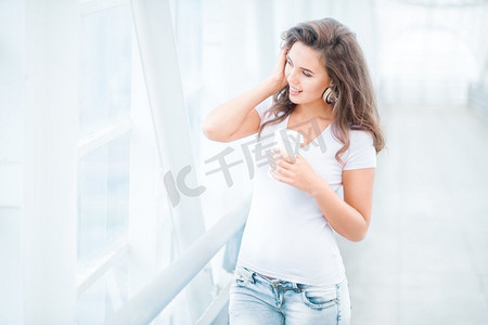 快乐的年轻女子戴着音乐耳机，站在桥上，手里拿着一个外卖咖啡杯，在城市背景的映衬下望向一边。