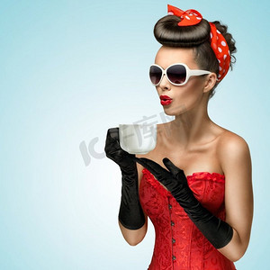 四分之三的照片中，迷人的美女戴着复古手套，头发上别着红丝带，手里拿着一杯热咖啡或茶，然后冷却。