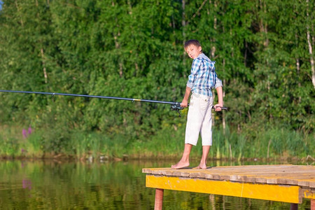 手里拿鱼摄影照片_穿着蓝色衬衫的男孩站在海边的码头上，手里拿着一根钓鱼竿