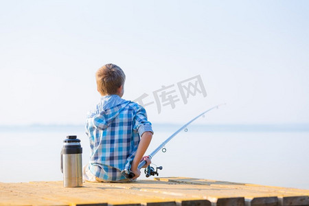 坐在海边摄影照片_穿蓝衬衫的男孩坐在馅饼上。穿着蓝色衬衫的男孩坐在海边的码头上，手里拿着钓鱼竿