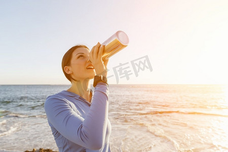 一名妇女在海滩上喝水。年轻女子在海滩上运动后喝水