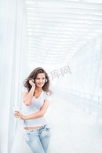快乐的年轻女子戴着音乐耳机，站在桥上，手里拿着一个外卖咖啡杯，在城市背景的映衬下望向一边。