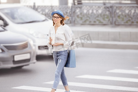 快乐的年轻女学生拿着咖啡外卖过马路，在城市的背景下开心地笑着。