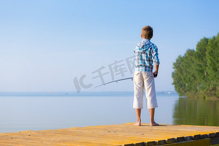 穿着蓝色衬衫的男孩站在海边的码头上，手里拿着钓鱼竿。穿蓝衬衫的男孩站在馅饼上
