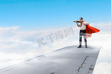 在飞行飞机的机翼上。年轻商人超级英雄在飞机机翼边缘拉小提琴