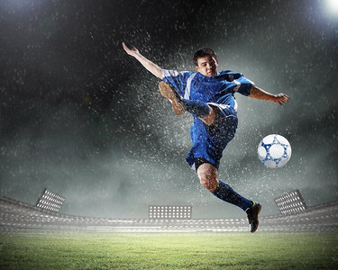人在雨中摄影照片_足球运动员击球。一个穿着蓝色球衣的足球运动员在雨中把球踢到了体育场的高处