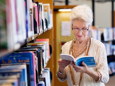 从容地摄影照片_一位老太太站在图书馆的书架旁。从容地读新书