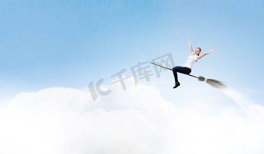 巫师摄影照片_穿着休闲的少女骑着扫帚在高空飞翔。扫帚上的女孩