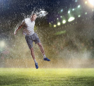 人在雨中摄影照片_足球运动员击球。在雨中的体育场用头击球的白色衬衫的足球运动员