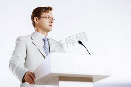 白色舞台摄影照片_一位年轻的商人站在舞台上对着麦克风讲话。舞台上的演讲者