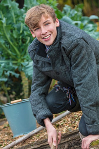 花园菜地摄影照片_快乐微笑的男孩子十几岁的年轻人在菜地里用水桶和花园叉子园艺