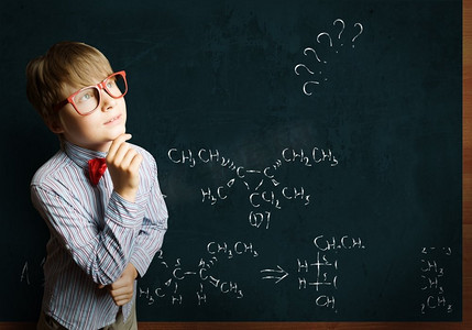 黑板公式摄影照片_聪明的小学生。戴着红眼镜的聪明男孩在黑板上写着公式
