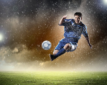 足球运动员击球。一个穿着蓝色球衣的足球运动员在雨中把球踢到了体育场的高处