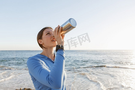 一名妇女在海滩上喝水。年轻女子在海滩上运动后喝水
